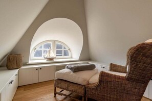A cozy attic bedroom with a sloped ceiling, a large arched window, a wicker chair, and a small table with a decorative item. - Holiday home in Borgsum with swimming pool (Borgsum)