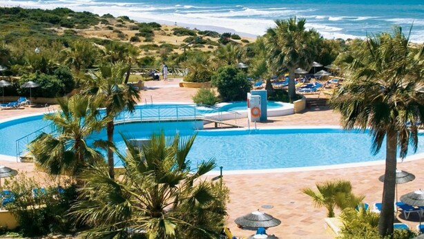 A resort pool area with a view of the ocean, surrounded by palm trees and lounge chairs. - Aldiana Club Andalusien (Chiclana de la Frontera)