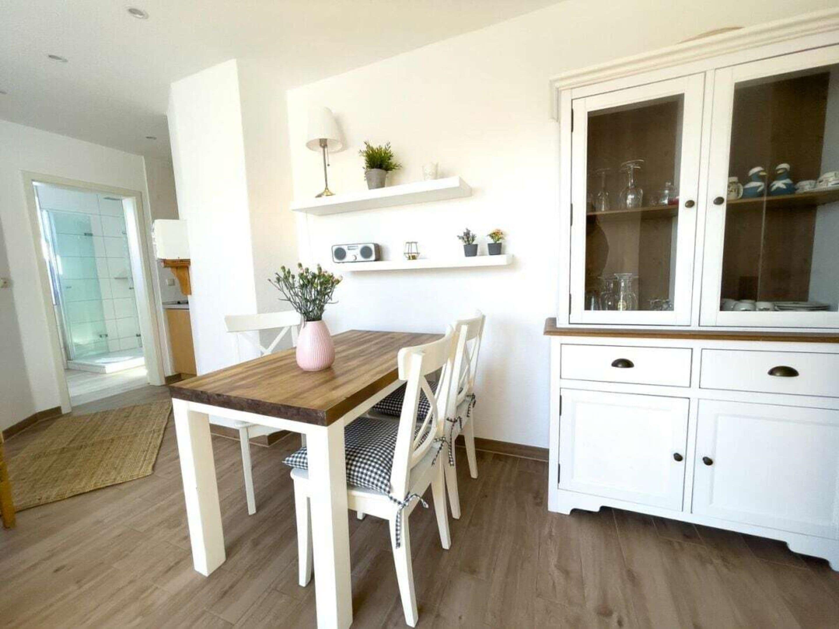 Un comedor con una mesa de madera y sillas blancas, un armario blanco con puertas de cristal y un pequeño estante con objetos decorativos.