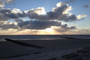 Una playa con un paseo marítimo pavimentado que conduce al agua, un cielo despejado con nubes, y el sol poniéndose o saliendo.