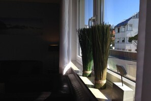A room with a window, a black leather sofa, and a tall green plant in the corner. - Holiday apartment with 1 bedroom (Norderney)