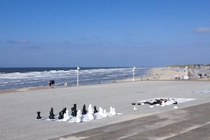 Een strand met een schaakbord in het zand, enkele mensen wandelen en de oceaan op de achtergrond.