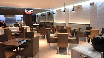 A modern restaurant interior with multiple tables and chairs, a television mounted on the wall, and pendant lighting.