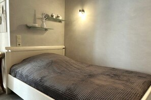 A single bed with a textured gray blanket, a wall-mounted shelf with decorative items, and a wall-mounted light fixture. - Holiday apartment with 1 bedroom (Norderney)