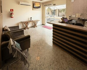 Zona de recepción de un hotel con mostrador, extintor, una pequeña mesa con una vela y un sofá con una revista.