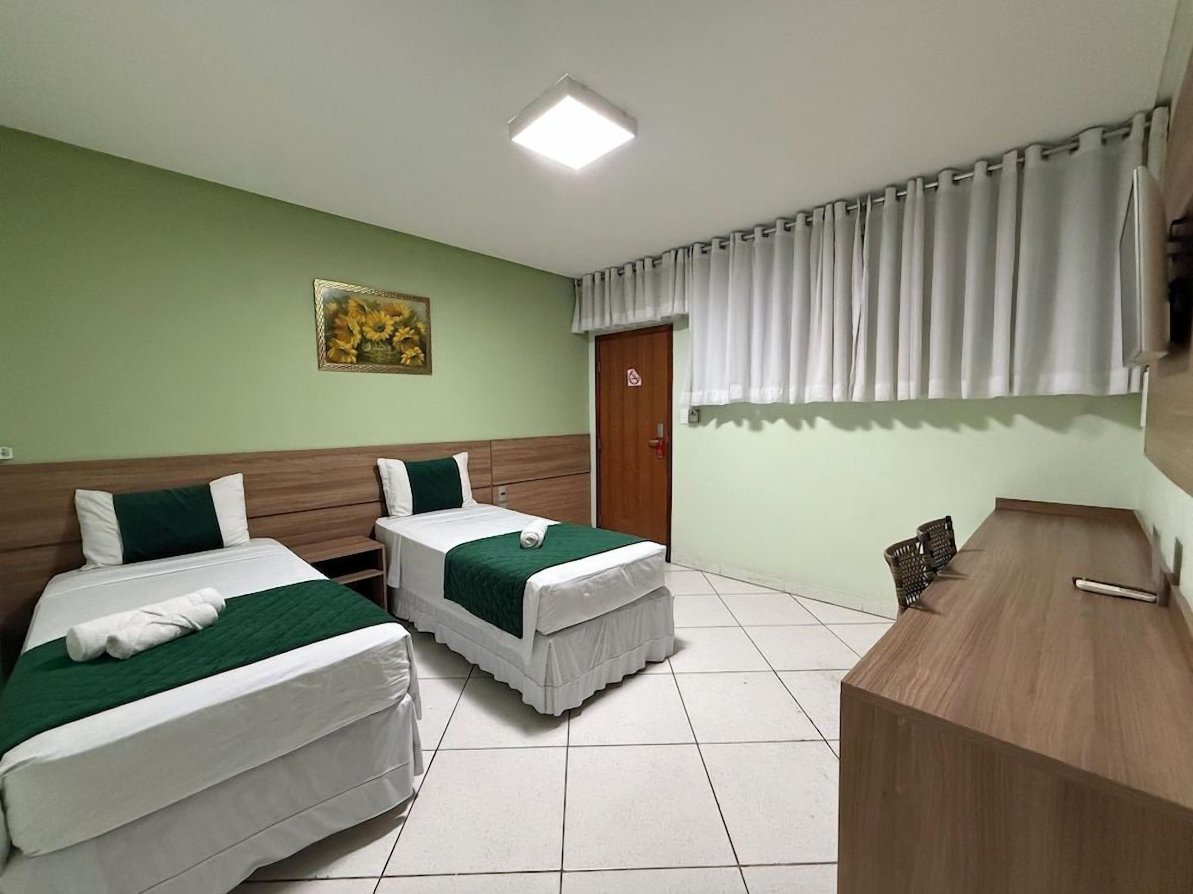 Quarto de hotel com duas camas, uma escrivaninha, janela com cortinas e um quadro na parede.