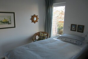 Ein Schlafzimmer mit einem Bett, einem Sessel, einem Fenster mit Vorhängen und einem Wandspiegel.