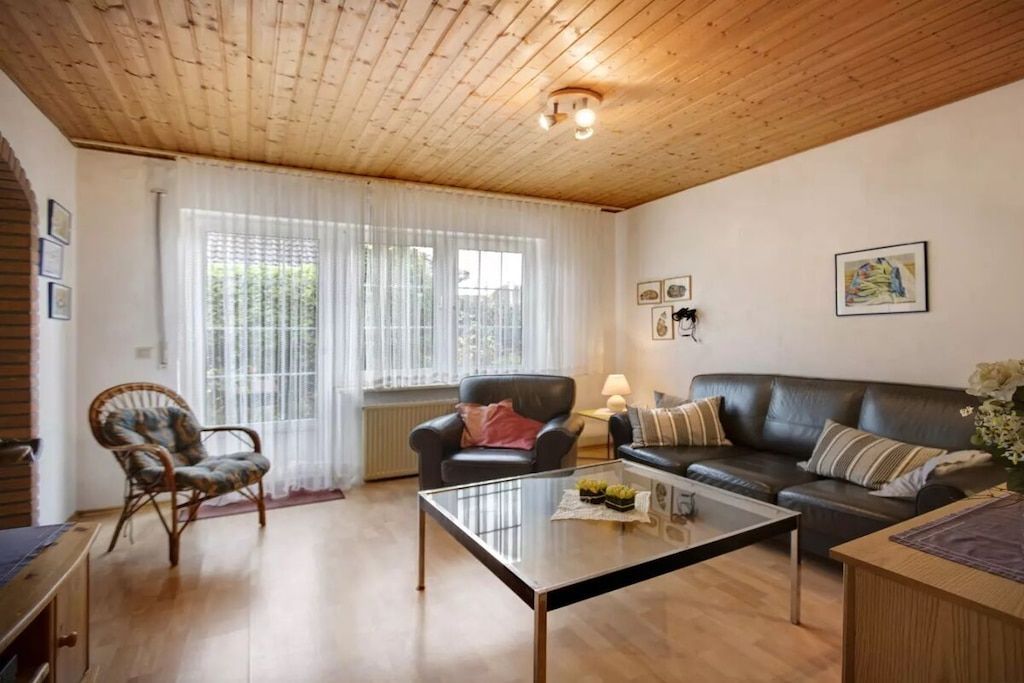 Un salon avec un plafond en bois, une table basse en verre, un canapé en cuir noir, un fauteuil en osier et une fenêtre ornée de rideaux légers.