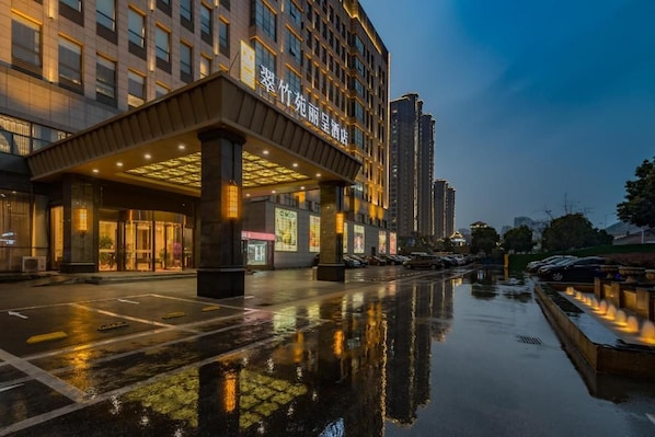 Hotel com entrada coberta, estacionamento e reflexo no chão molhado.