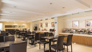Uma sala de jantar moderna com várias mesas e cadeiras, área de cozinha e balcão com distribuidores de alimentos.