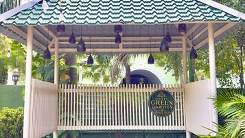 Um gazebo com telhado verde, uma placa com a inscrição 'Hotel Green Garden' e uma área de entrada pavimentada com um pequeno jardim.
