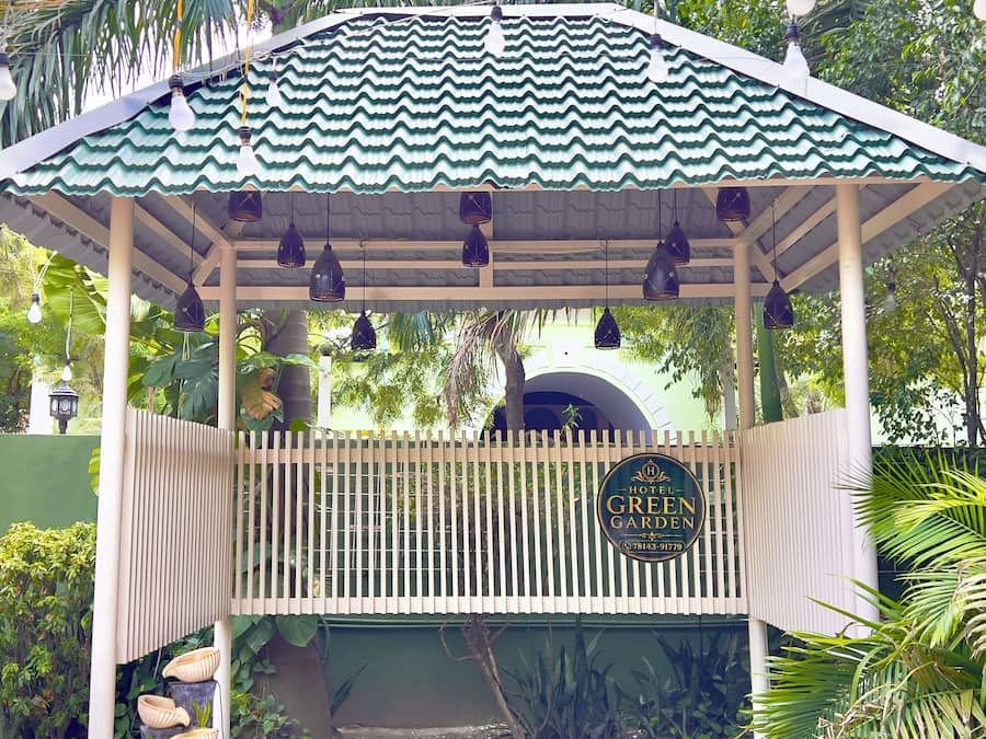Un belvédère surmonté d’un toit végétalisé, une enseigne indiquant « Hôtel Green Garden », et une zone d’entrée pavée agrémentée d’un petit jardin.