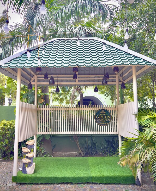 A gazebo with a green roof, a sign reading 'Hotel Green Garden,' and a paved entrance area with a small garden.