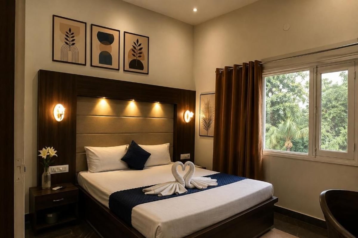 A hotel room with a large bed, a nightstand with a vase of flowers, and a view of greenery through the window.