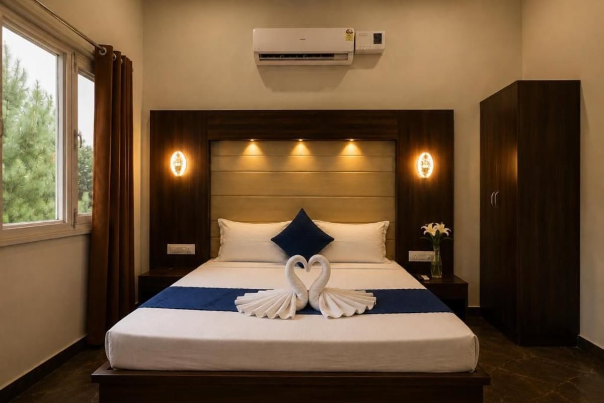 A hotel room with a large bed, a headboard with integrated lighting, an air conditioning unit, and a view of trees through the window.