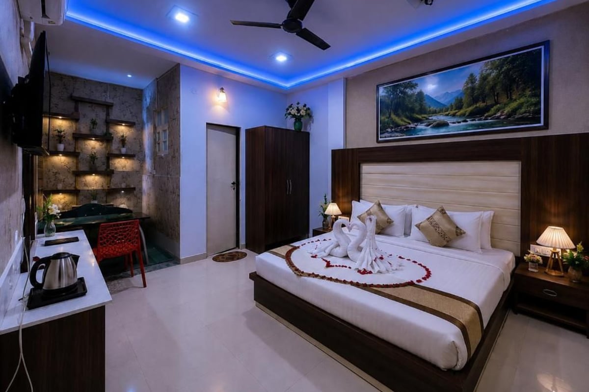 A hotel room with a bed, a desk, a chair, a TV, a ceiling fan, and a painting.