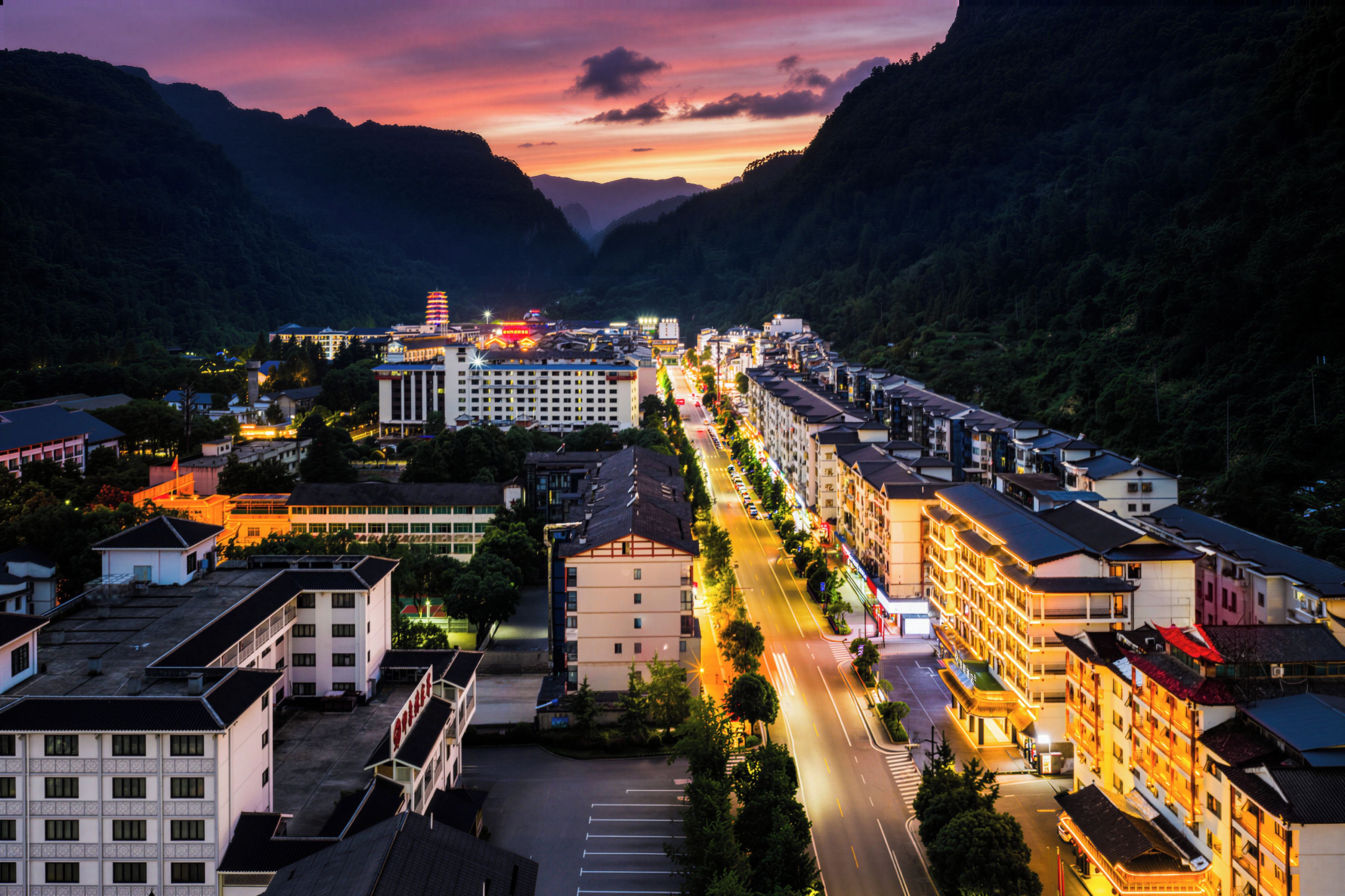 A town with a long street lined with buildings, a prominent hotel with a tower, and mountains in the background.