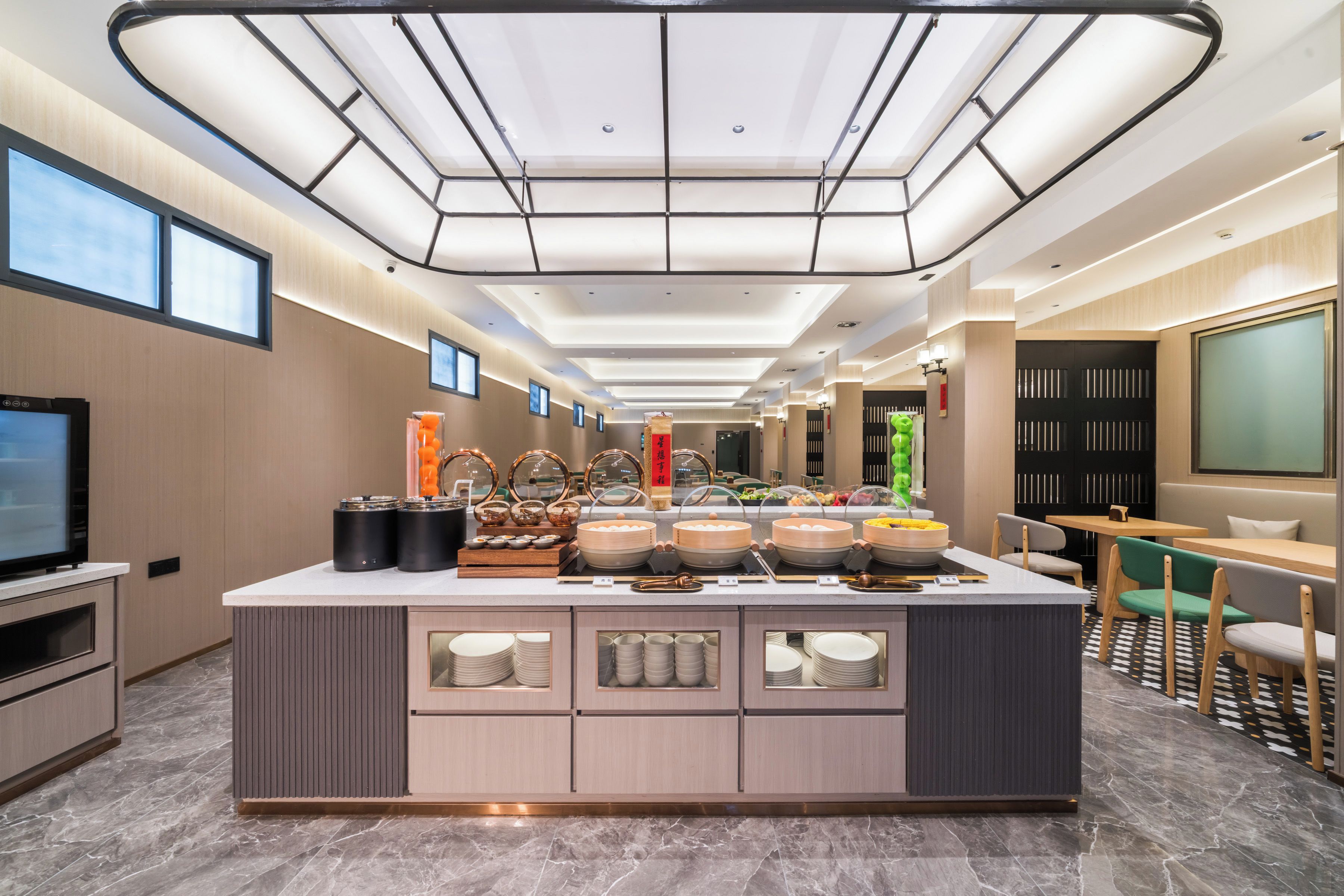 A modern hotel lobby with a central buffet area, a television, and a seating area with tables and chairs.