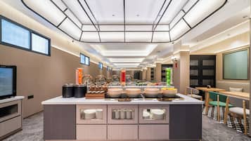 A modern hotel lobby with a central buffet area, a television, and a seating area with tables and chairs.