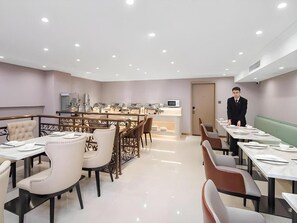 En restaurant i et hotel med flere borde og stole, et buffetbord og en medarbejder.