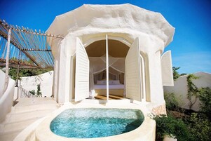 Una estructura blanca, de ocho lados, con un acceso abierto que da a una cama con sábanas blancas. Frente a la estructura, una bañera de hidromasaje circular.