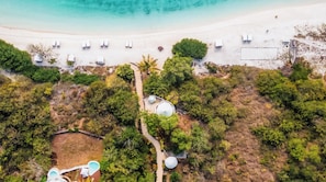 Ett flygbildsfoto av en strand med vit sand, en stig och flera vita byggnader, troligen vattentankar, omgivna av grönska.