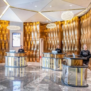 Ein Hotel-Empfangsbereich mit drei Empfangstresen, einem modernen Innenraum mit metallischen und hölzernen Elementen sowie einer großen digitalen Anzeige an der Wand.