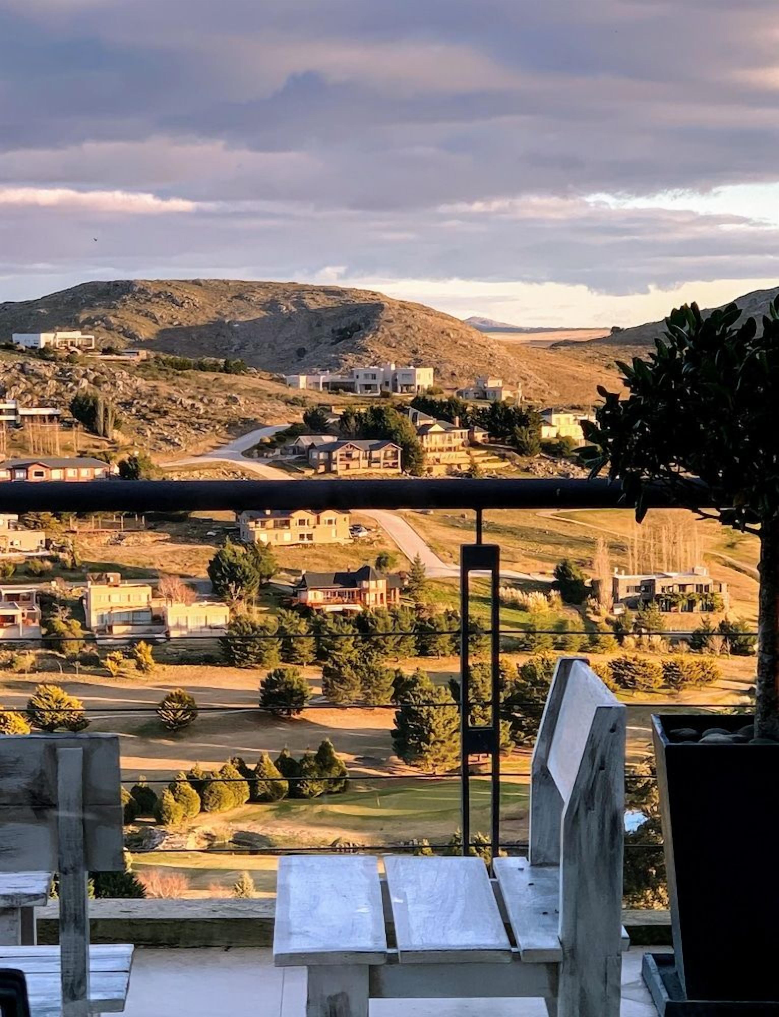 A view from a balcony overlooking a residential area with houses, a road, and hills in the distance.