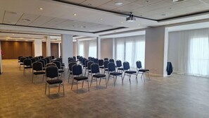 Un salón de conferencias con filas de sillas, un proyector y ventanas amplias.