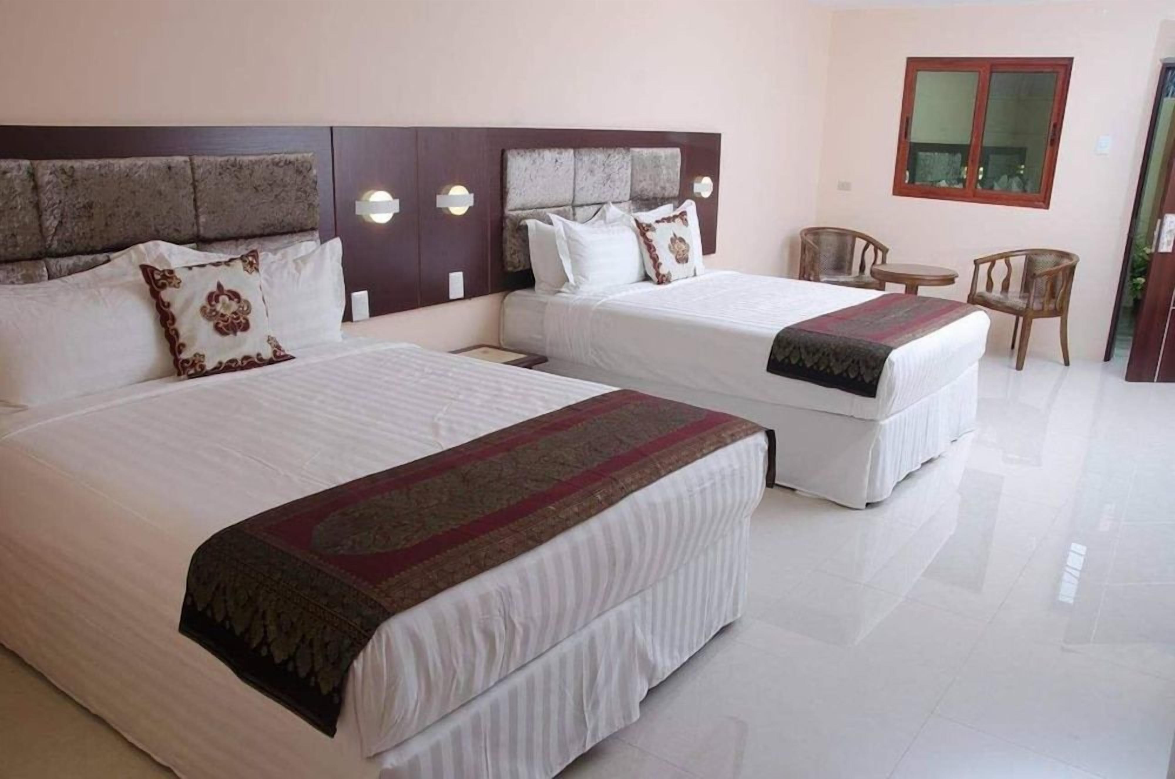A hotel room with two beds, a wooden headboard, a small table with chairs, and a window.