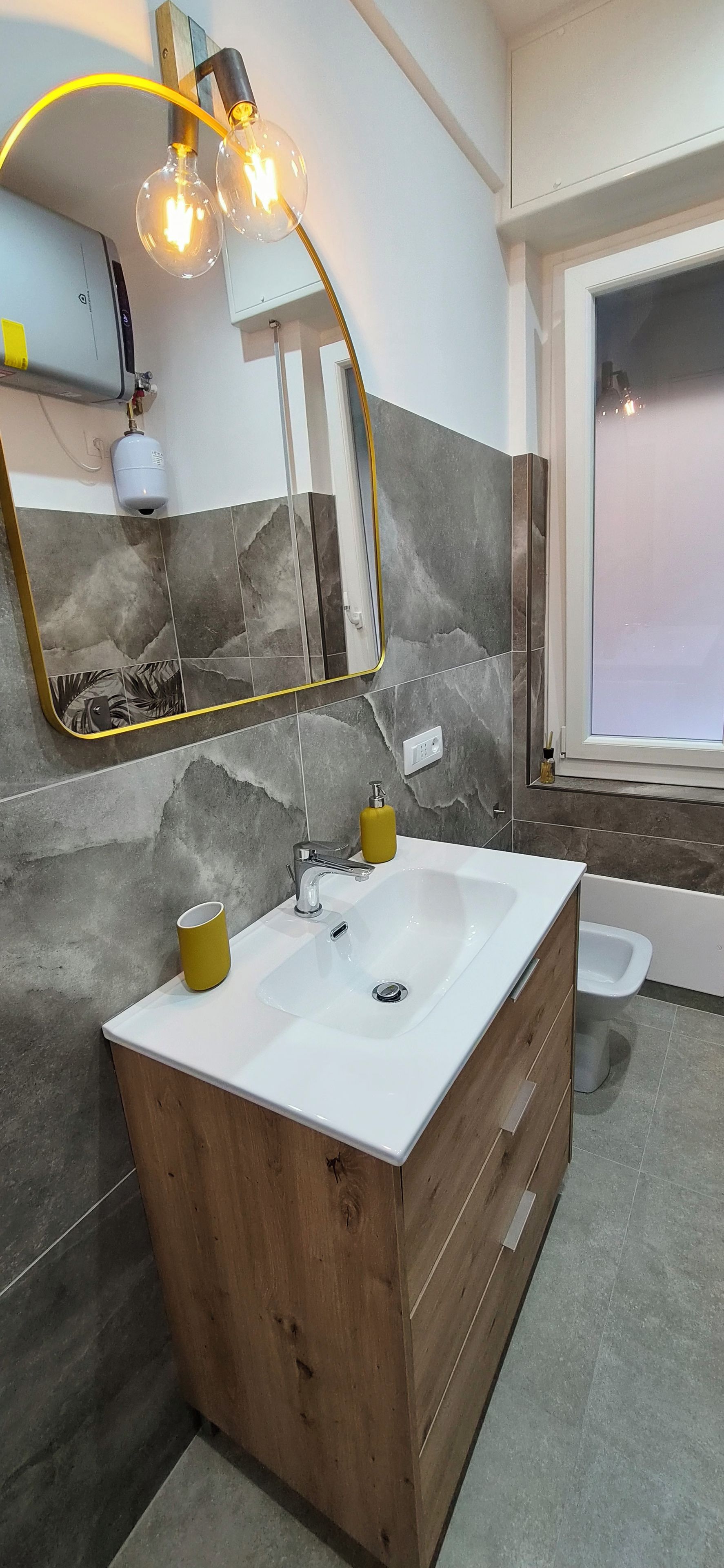 Ein modernes Badezimmer mit einem Holz-Waschtisch, einem weißen Waschbecken, einem Spiegel mit goldener Rahmung und einem Fenster.