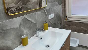 Ein modernes Badezimmer mit einem Holz-Waschtisch, einem weißen Waschbecken, einem Spiegel mit goldener Rahmung und einem Fenster.