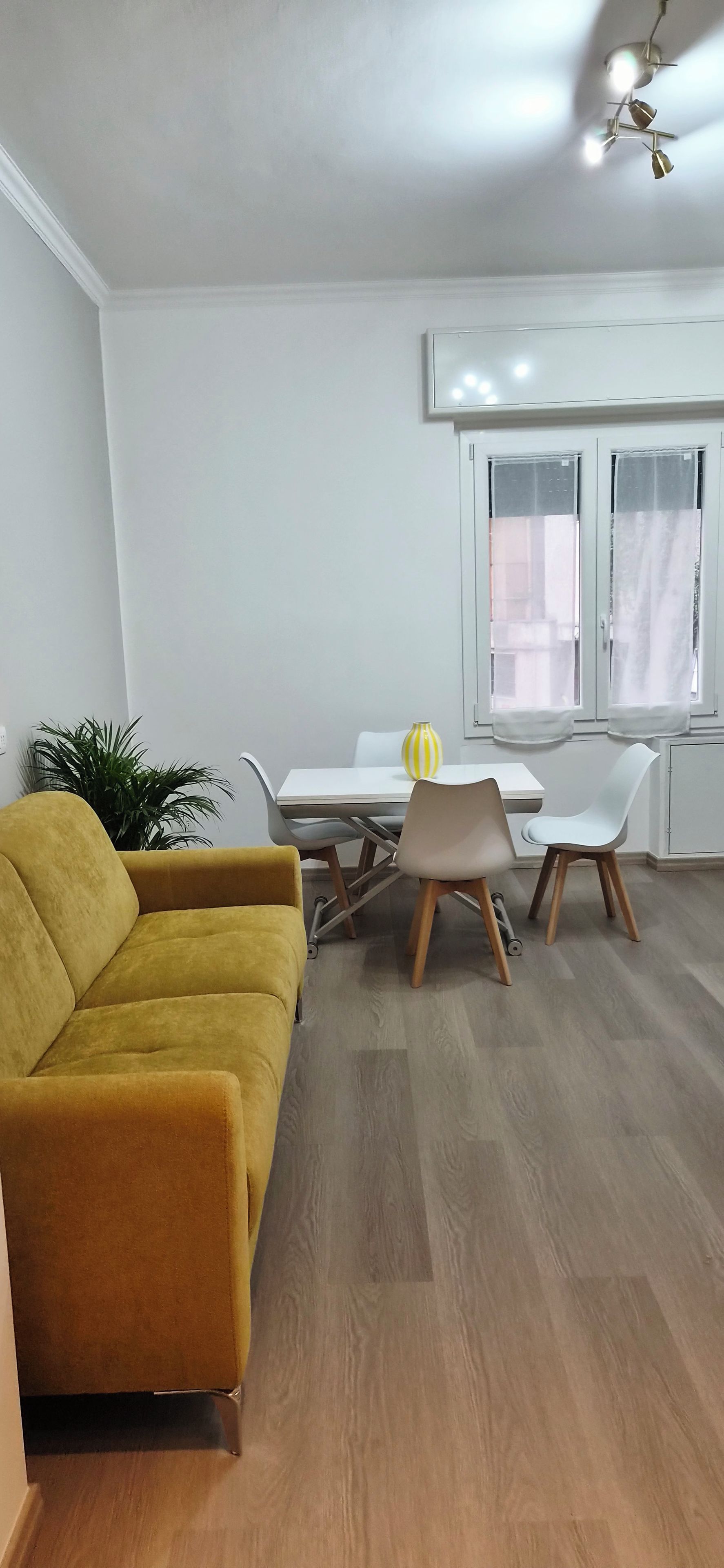 Ein modernes Wohnzimmer mit einem gelben Sofa, einem Esstisch mit Stühlen und einer Zimmerpflanze.