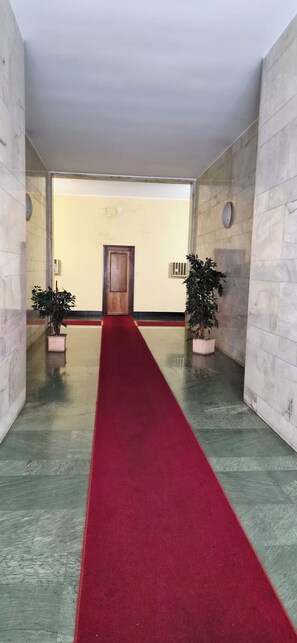 A hallway with a red carpet leading to a door, flanked by potted plants and marble walls.