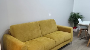 Quarto com sofá amarelo, mesa branca e planta em vaso.