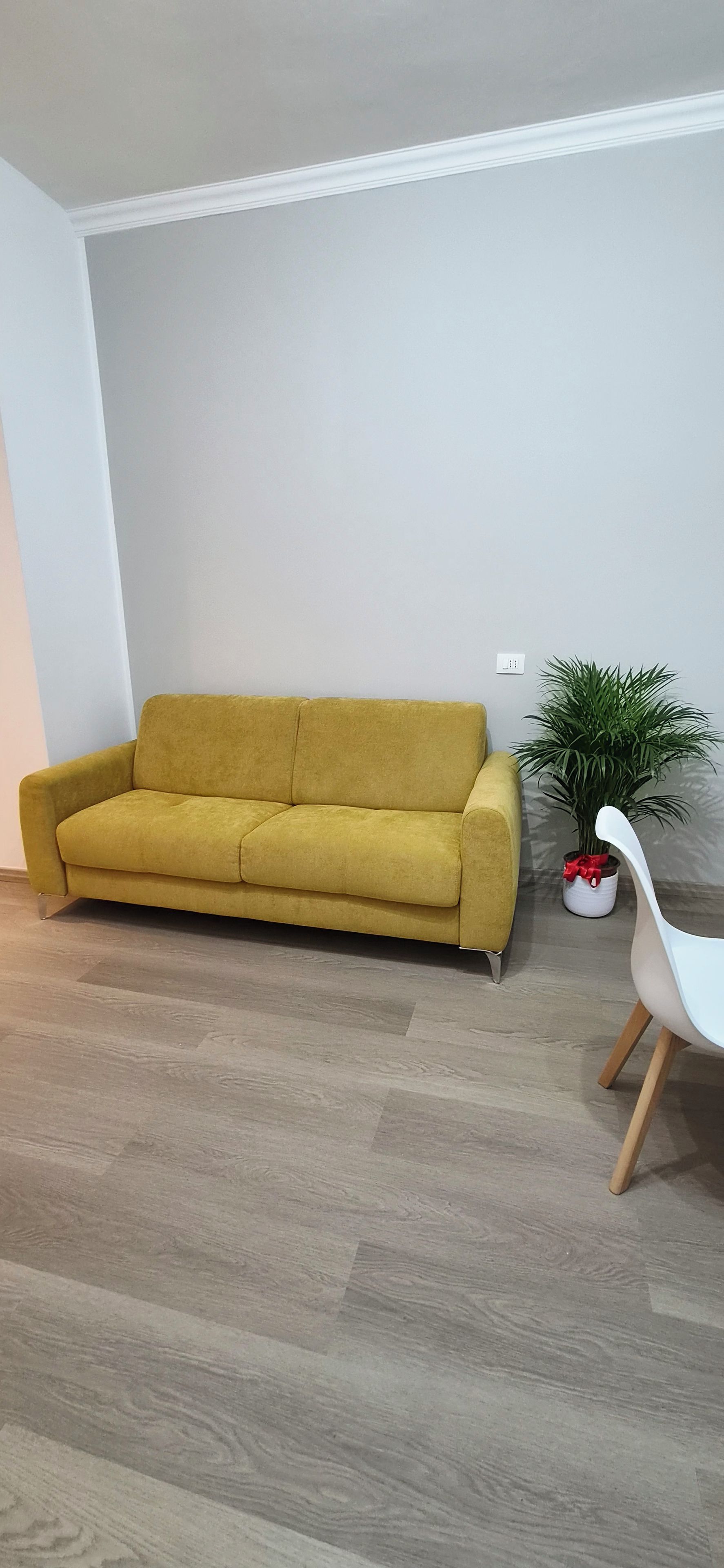 Ein Zimmer mit einem gelben Sofa, einem weißen Sessel, einer Topfpflanze und Holzboden.