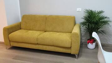 Ein Zimmer mit einem gelben Sofa, einem weißen Sessel, einer Topfpflanze und Holzboden.
