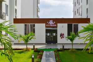 Ein Fitnessstudio im modernen Design mit Holzfassade, Glastür und einem gepflegten Garten mit Palmen und Blumen.