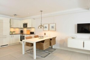 A modern kitchen with white cabinetry, a dining table with chairs, a pendant light, and a flat-screen TV on the wall. - Holiday apartment in Sylt with 2 bedrooms (Sylt \/ Westerland)