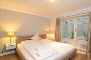 A hotel room with a double bed, two bedside lamps, a window with curtains, and a bedside table. - Holiday apartment in Sylt with 2 bedrooms (Sylt \/ Westerland)