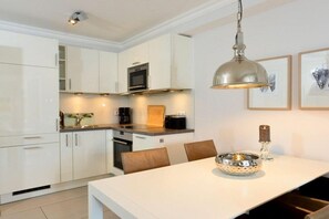 A modern kitchen with white cabinetry, a stainless steel range hood, and a dining area with a white table and brown chairs. - Holiday apartment in Sylt with 2 bedrooms (Sylt \/ Westerland)