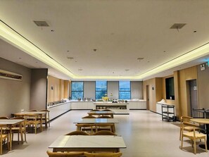 Un amplio comedor de hotel con mesas y sillas largas, una barra central y ventanas grandes que dejan entrar luz natural.