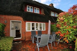 A brick house with a thatched roof, a dining area with chairs and a table, and a small garden with a bush. - in the pastorate courtyard, house 3 in Ohl Dörp 22 (Wrixum)