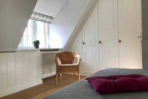 Ein Schlafzimmer mit schrägen Wänden, einem Fenster mit Pflanze, einem Korbstuhl und einem Bett mit gemustertem Überwurf und einem violetten Kopfkissen.