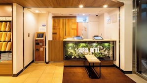 Zona de recepción de un hotel con un mostrador de madera, un quiosco digital de registro y un letrero que dice 'Super Hotel'.