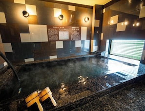 A hot spring bath with wooden stools, a tiled wall with informational plaques, and a view of the outside through a window. - Super Hotel Totsuka Eki Higashiguchi (Yokohama)