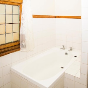 A bathroom with a bathtub, a window with a wooden frame, and white tiled walls. - Jikeleza Lodge Backpackers (Gqeberha)