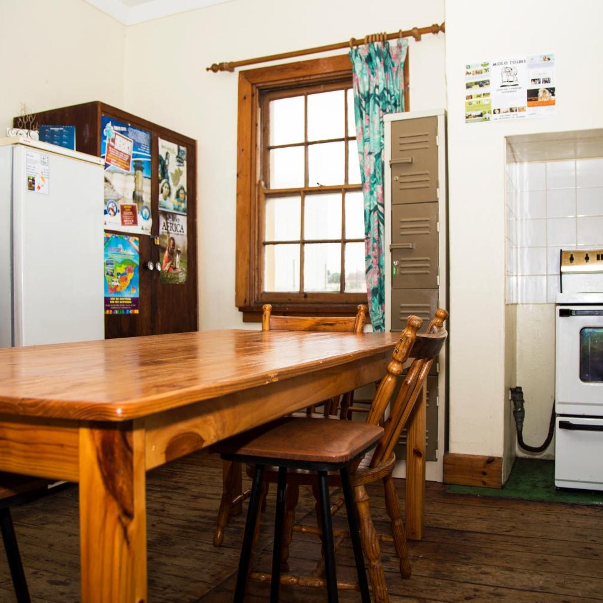 A kitchen with a wooden table and chairs, a window with curtains, a refrigerator, and a wall with various notices.