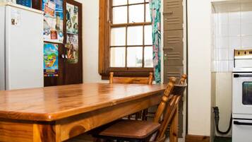 A kitchen with a wooden table and chairs, a window with curtains, a refrigerator, and a wall with various notices.