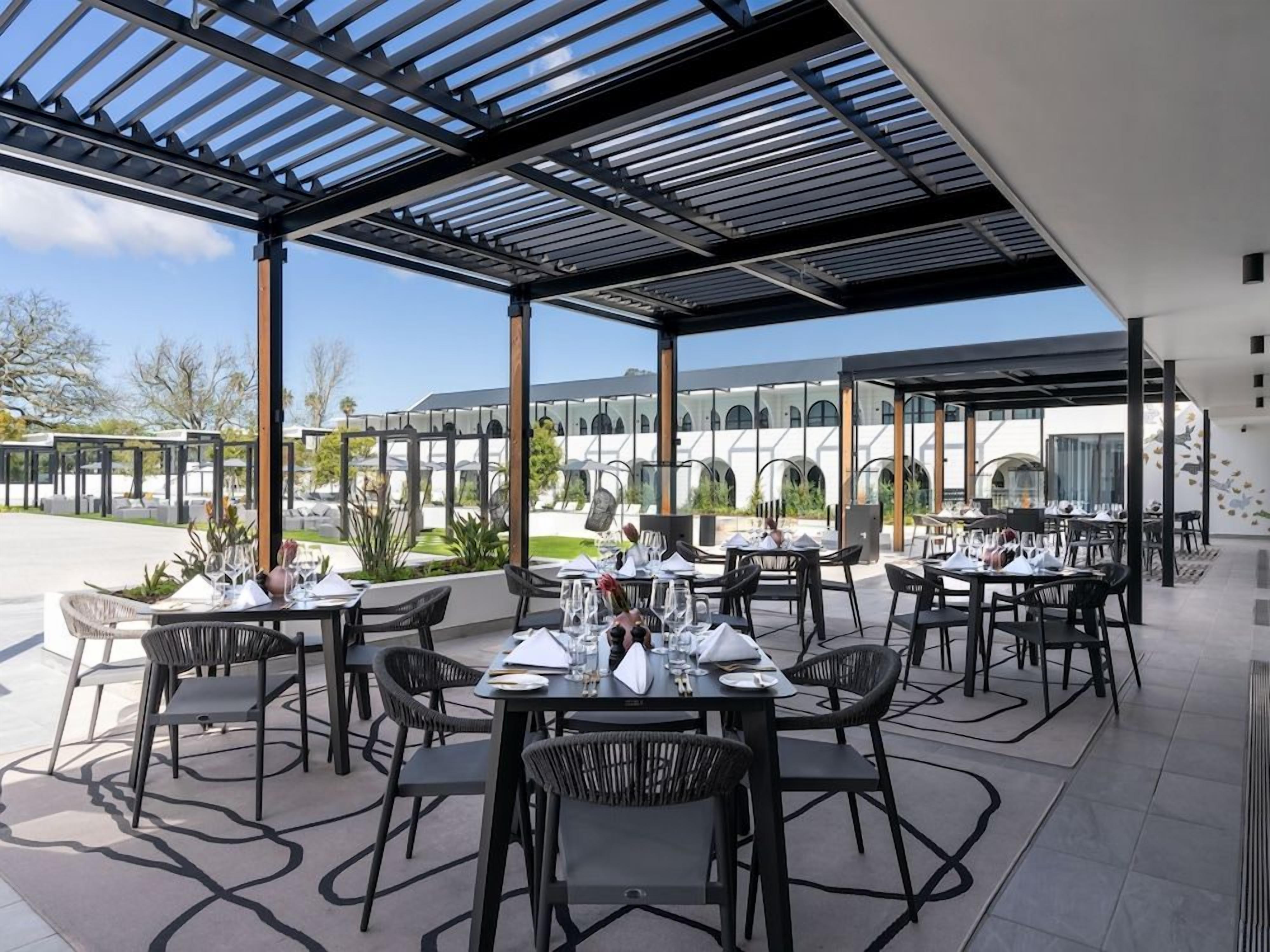 An outdoor dining area with tables and chairs, a pergola structure, and a view of a building with arches in the background.